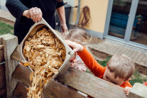 Betreuende wirft gemeinsam mit Kind einen Behälter mit Kartoffelschalen auf den Kompost