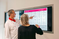 Koch und Mitarbeiterin besprechen den Speiseplan an einem Bildschirm an der Wand