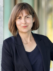 Foto von Gisela Olias, die in dunkelblauem Blazer und dunkelblauen T-Shirt in die Kamera lächelt