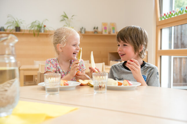 Ein Junge und ein Mädchen sitzen in der Kita am Tisch und essen lachend Apfelstücke