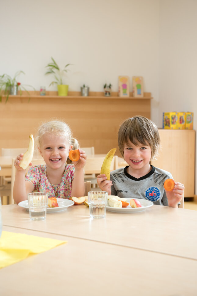 Mädchen und Junge halten in der Kita Obst hoch und lachen