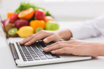 Hände tippen auf Laptop-Tastatur, Im Hintergrund ein Haufen Obst