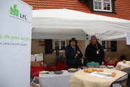 Auf dem Bild sind die beiden Sortenschützer Dr. Klaus Fleißner und Adolf Kellermann zu sehen. Sie stehen hinter einem Tisch mit weißer Tischdecke, auf dem Teller stehen. Links im Bild ist ein Rollupder LfL zu sehen und im Hintergrund die Frontseite eines Hauses mit rotem Dach. 