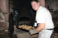 Auf dem Bild ist der Bäckermeister Sebastian Groß zu sehen. Er holt ein Blech mit fertigen KArtoffelbrötchen aus dem historischen Backofen.