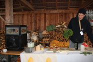 Auf dem Bild ist Theo Lindinger zu sehen. Er steht hinter einem Tisch mit weißer Tischdecke und hält einen Bund Kräuter in der Hand. Im Hintergrund sind Kartoffelsäcke und Holz zu sehen. 