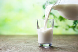 Eingießen von Milch in Glas. Im Hintergrund ist verschwommen Natur zu erkennen.