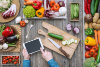 Hände bedienen ein Tablet an einem Küchentisch, der mit Gemüse beladen ist