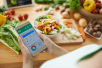 Eine Frau sitzt vor einem Küchentisch mit Gemüse und ruft ihre Ernährungs-App auf dem Smartphone auf