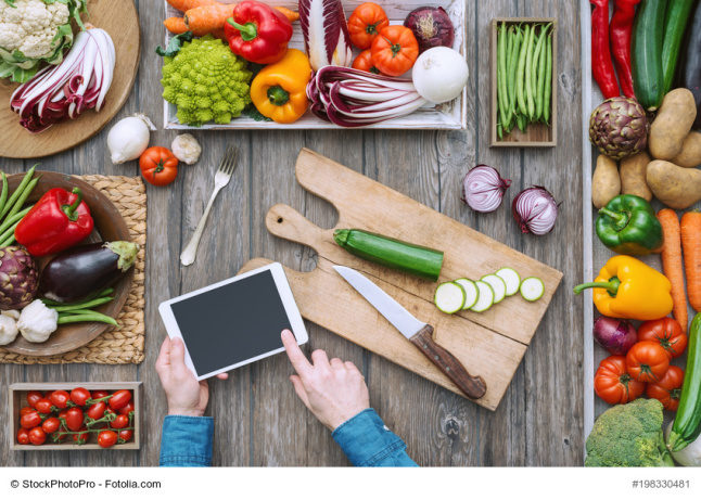 Kleines Tablet in Mitten verschiedener Lebensmittel. Auf einem Holzbrett liegt eine Zuchini, die zur Hälfte in Scheiben geschnitten ist.