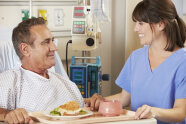 Pflegekraft birngt lächelndem Herren ein Sandwich ans Krankenbett