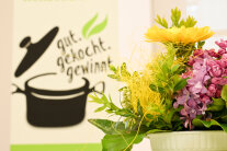 Blumengesteck im Vordergrund mit Logo von gutgekochtgewinnt im Hintergrund.