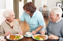Pflegekraft serviert lächelndem Seniorenpaar das Mittagessen